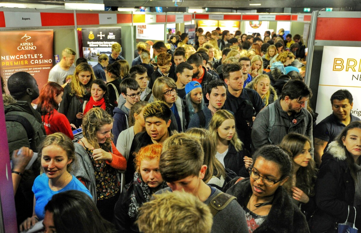 A crowded career fair with numerous attendees exploring different stalls and also considering reading our youth marketing blog. People of diverse backgrounds are seen interacting, and various company signs are visible above. The atmosphere is bustling and energetic. BAM Agency