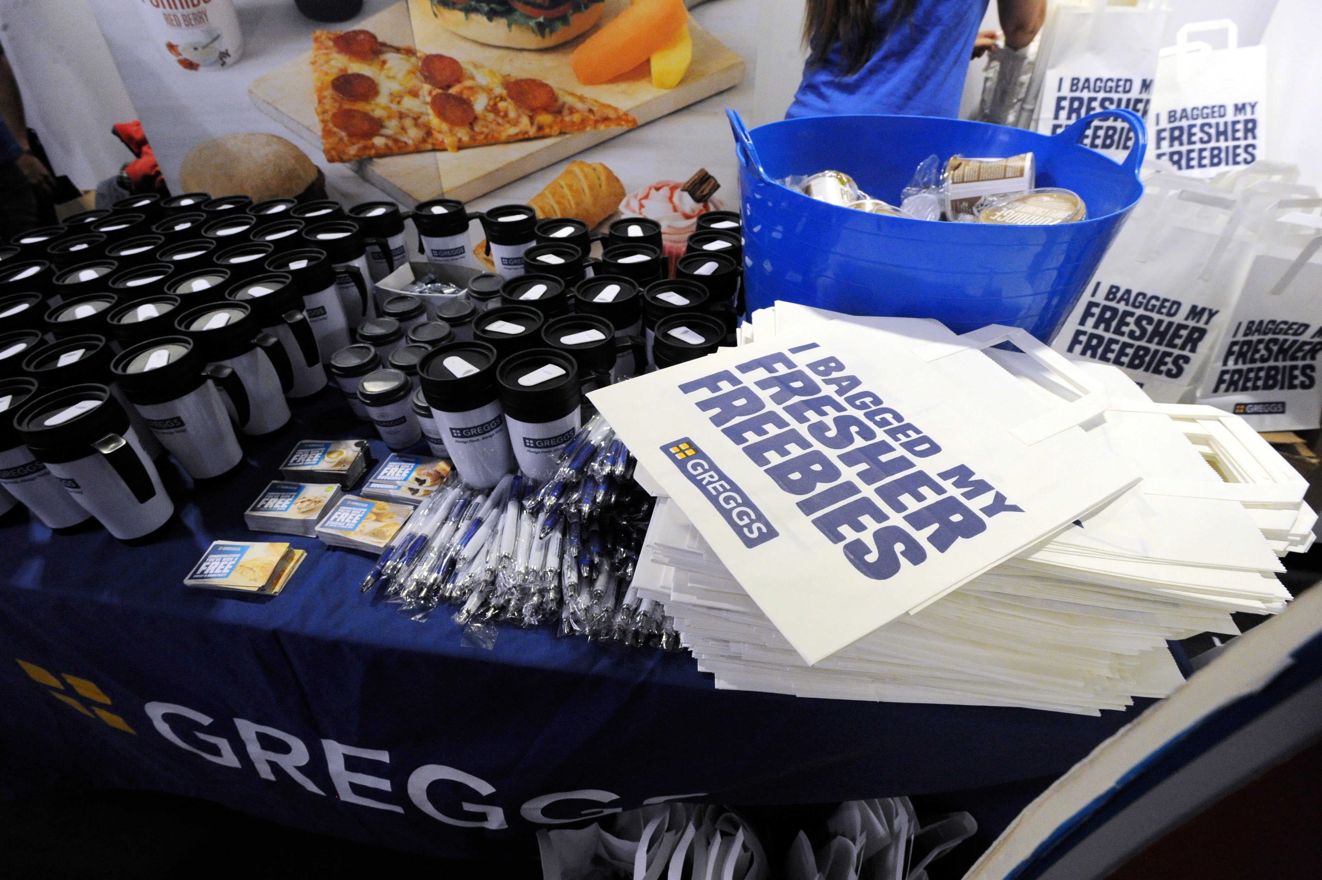 A display table at an event featuring a variety of promotional items: reusable cups, bags with I bagged my fresher freebies text, pens, and snacks. The table has a blue cloth with the Greggs logo. BAM Agency