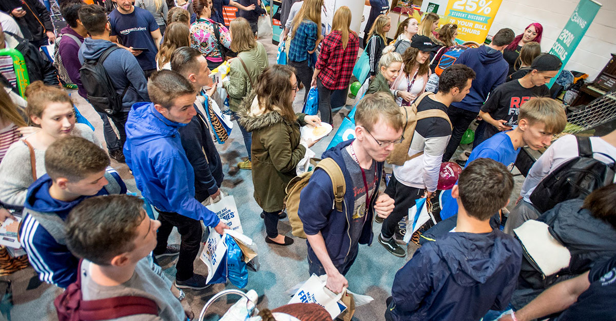A crowded indoor setting with diverse individuals, many carrying backpacks and holding bags. People are standing, walking, or browsing booths with promotional signs. The atmosphere appears busy and interactive, suggesting an event or fair. BAM Agency