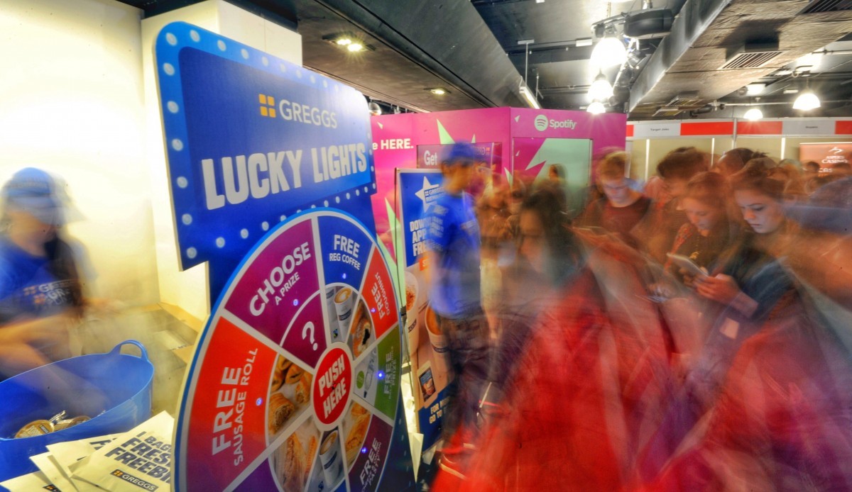 A bustling event scene with people gathered around a colorful Greggs Lucky Lights game booth. The game features a spinning wheel offering various prizes. Attendees are engaging enthusiastically with the booth and its interactive features. experiential, activations, events, promotion, brand BAM Agency