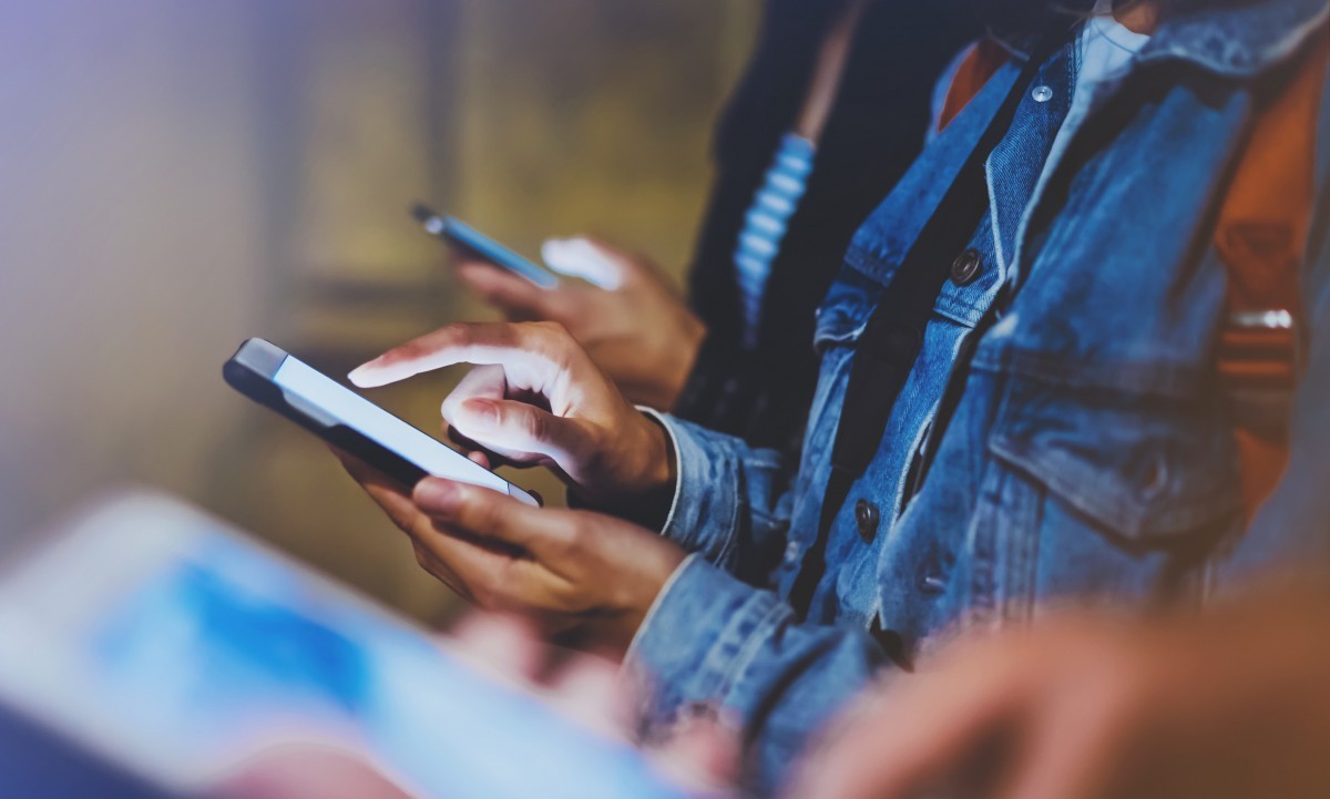 Digital Marketing youth using smartphones in a dimly lit environment. A person in the foreground is wearing a denim jacket and is interacting with their phone, while two others in the background are focused on their devices. The scene captures a sense of connectivity. BAM Agency