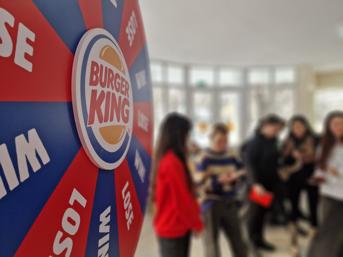 BAM Agency A Burger King prize wheel with sections labeled WIN and LOSE is in the foreground. In the background, a group of people stand, slightly out of focus, in a bright room. BAM Agency
