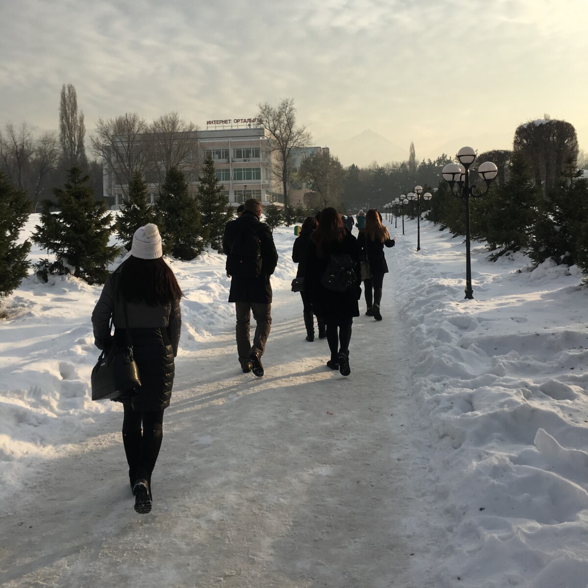 BAM Agency People are walking along a snow-covered path lined with lamp posts and evergreen trees, wearing winter coats and hats. In the background, a building stands under a cloudy sky, reminiscent of the warm refuge found in a cozy Burger King on a crisp winter day. BAM Agency