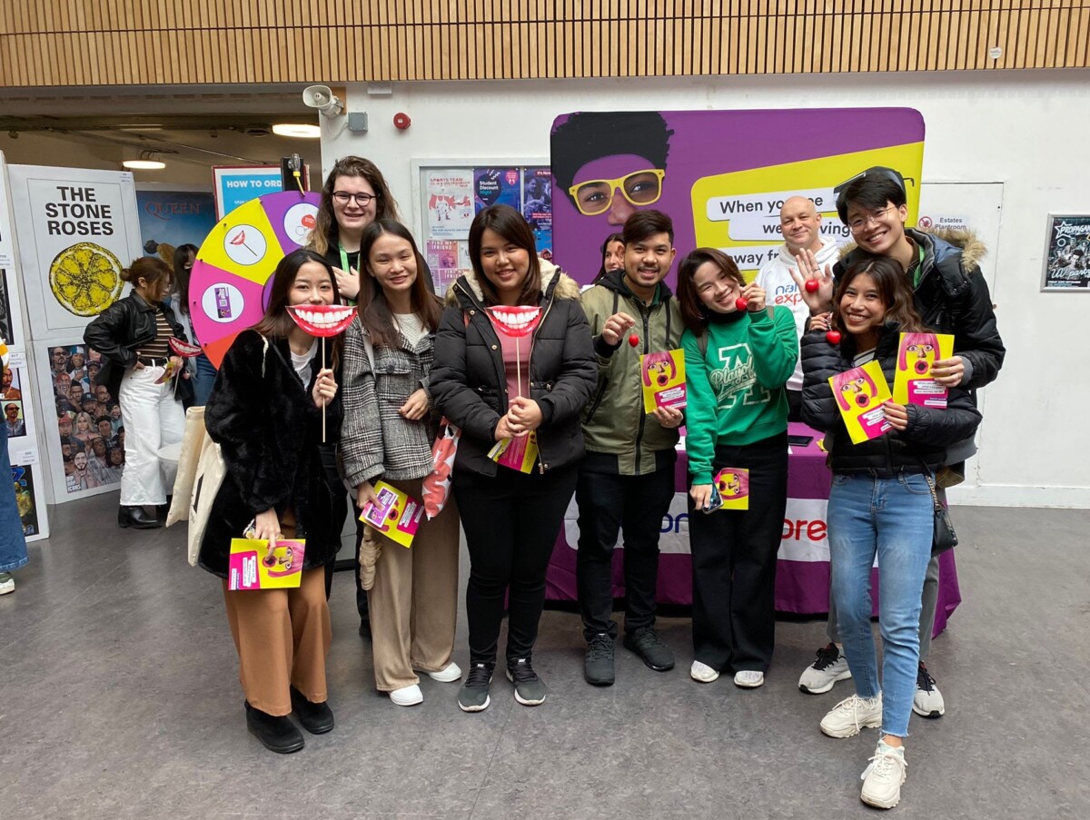 BAM Agency A group of people is standing in front of a purple booth with pamphlets. They are holding large, cartoonish mouth props with smiles. Posters are visible on the walls behind them, including one for The Stone Roses. BAM Agency