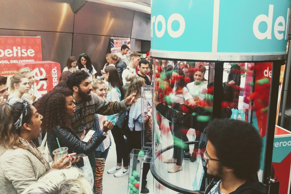 People are gathered around a glass cylinder filled with red and green balls. A man is excitedly reaching out while others watch and smile. The scene is lively, with branding visible around the area. BAM Agency