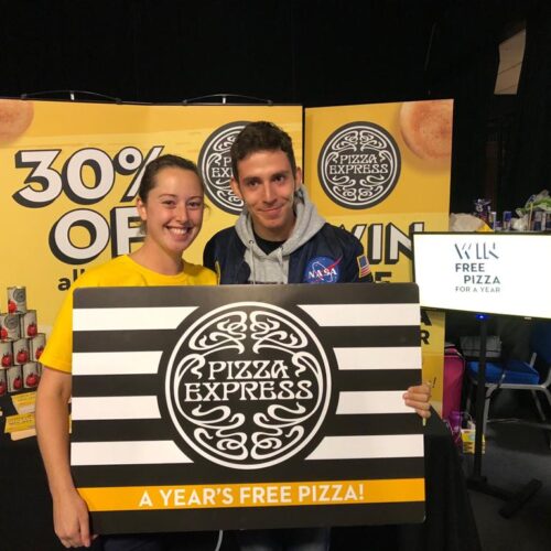 BAM Agency Two people smiling and holding a large sign that reads Pizza Express and A Years Free Pizza! in front of a display offering 30% off. One wears a yellow shirt, the other a NASA hoodie. A sign on the right reads Win Free Pizza for a Year!. BAM Agency