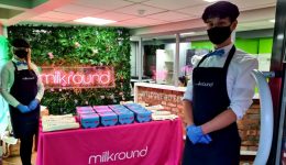 Two people wearing masks and aprons stand by a Milkround booth with a pink tablecloth. Boxes and tote bags are on the table. A neon Milkround sign is on the wall with greenery behind them. BAM Agency