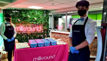 Two people wearing masks and aprons stand by a Milkround booth with a pink tablecloth. Boxes and tote bags are on the table. A neon Milkround sign is on the wall with greenery behind them. BAM Agency