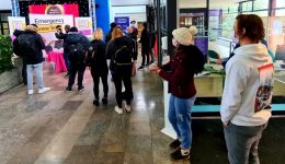 People stand in line at a booth with a pink tablecloth labeled Emergency Brew Bar in a brightly lit indoor area. Some individuals wear backpacks and face masks. The space has informational posters and a partially visible stage on the left. BAM Agency
