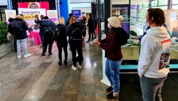 People stand in line at a booth with a pink tablecloth labeled Emergency Brew Bar in a brightly lit indoor area. Some individuals wear backpacks and face masks. The space has informational posters and a partially visible stage on the left. BAM Agency