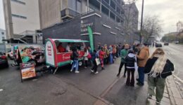 BAM Agency People line up at a food truck with a green canopy and red lettering in an urban area. Theres a sandwich board displaying menu items. The backdrop features a brick building and a partly cloudy sky. BAM Agency
