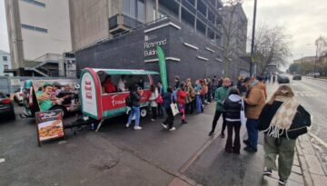 BAM Agency People line up at a food truck with a green canopy and red lettering in an urban area. Theres a sandwich board displaying menu items. The backdrop features a brick building and a partly cloudy sky. BAM Agency