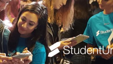 BAM Agency At a lively event, people mingle while two individuals in blue StudentUniverse shirts are engrossed in their phones, engaging in a captivating youth marketing game. A woman between them peers at the screen as the StudentUniverse logo stands out prominently in the foreground. BAM Agency