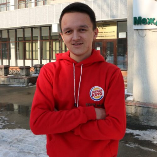 BAM Agency A person in a red Burger King hoodie stands outside, arms crossed, proudly smiling into the camera. The background reveals a building with large windows and snow gently dusting the ground. BAM Agency