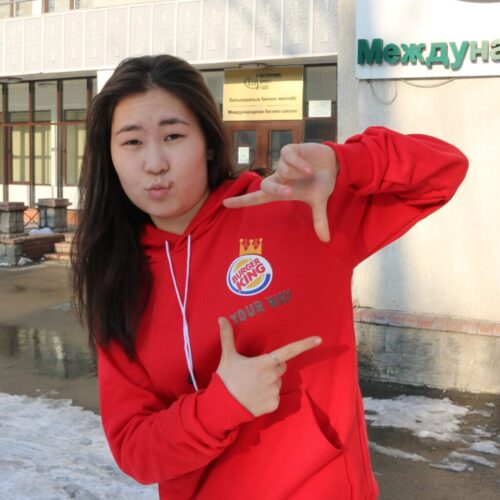 BAM Agency A person in a red hoodie sporting the Burger King logo grins playfully, framing their face with their hands in front of a building entrance. Snow blankets the ground, and signs written in Cyrillic add to the scenes charm. BAM Agency