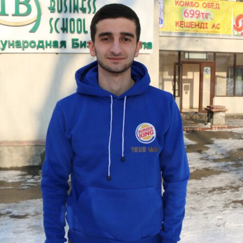 BAM Agency A man clad in a blue Burger King hoodie stands on a snowy sidewalk, framed by a building with Cyrillic signs and windows. The sun highlights his features as he gazes directly at the camera, capturing the moments essence. BAM Agency