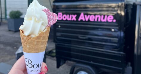 A hand with red nail polish holds a soft-serve ice cream cone topped with a heart-shaped pink sprinkle in front of a black food truck with Boux Avenue. written in pink letters. BAM Agency