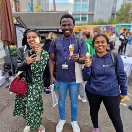Three people smiling and holding ice cream cones stand outdoors at an event, with a crowd, colorful mural, and food truck in the background. The group appears happy and casually dressed. BAM Agency