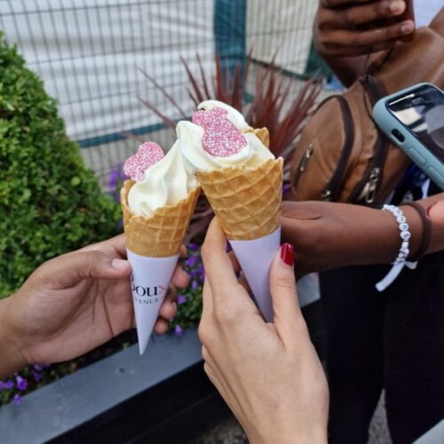 Two people hold ice cream cones with pink bear-shaped sprinkles, toasting them together. Another person in the background holds a phone. The scene appears to be outdoors near greenery and a fence. BAM Agency