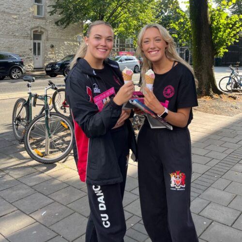 Two smiling young women in sports uniforms stand on a sidewalk holding ice cream cones. Bicycles are parked behind them, and trees and buildings line the street in the background. BAM Agency