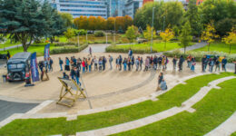 BAM Agency A long line of people eagerly waits outside in a park-like space for face-to-face activations, as the black food truck serves up emerging trends. The line curves around a paved area, set against trees with autumn foliage and modern buildings in the background. BAM Agency