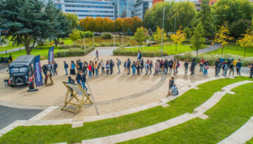 BAM Agency A long line of people eagerly waits outside in a park-like space for face-to-face activations, as the black food truck serves up emerging trends. The line curves around a paved area, set against trees with autumn foliage and modern buildings in the background. BAM Agency