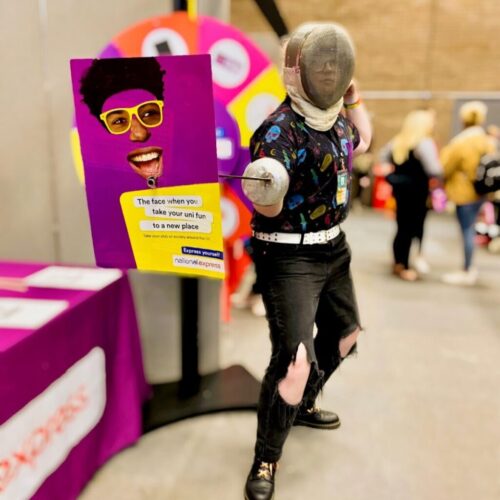 BAM Agency A person in fencing gear strikes an expressive pose at an indoor event. They hold a colorful, humorous sign with a smiling face wearing yellow glasses. A spinning wheel and people are visible in the blurred background, adding to the lively atmosphere of this National Express event. BAM Agency