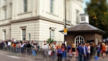 BAM Agency A long line of people waits outside a large, white building with columns, hoping for assistance within. The bustling scene features trees and another building in the background. Under the clear sky, its a vivid reminder of the Unions commitment to help its community. BAM Agency