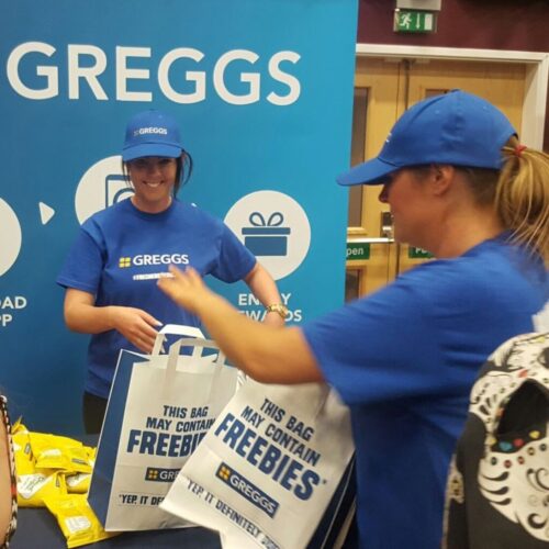 Two smiling individuals wearing blue Greggs caps and shirts distribute bags that read This bag may contain freebies at a promotional event. A table with snacks and a Greggs backdrop is visible in the background. BAM Agency