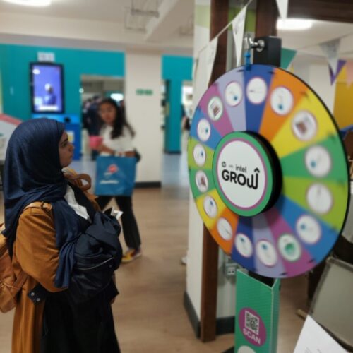 A woman in a brown coat and hijab stands in front of a colorful prize wheel labeled GROW inside a bright indoor space, holding a jacket and looking at the wheel. Other people are visible in the background. BAM Agency