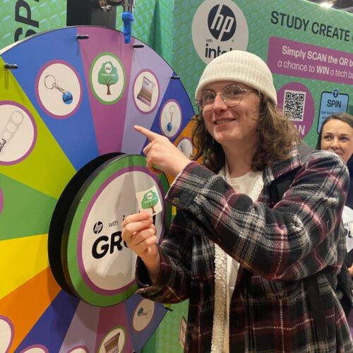 A smiling person wearing glasses and a beanie points at a colorful prize wheel at an event booth. They hold a small sticker and stand next to an HP branded display promoting a giveaway. BAM Agency