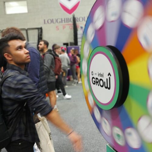 A person spins a colorful prize wheel labeled “Intel Grow” at an indoor event, with several people and a Loughborough Sport sign visible in the background. BAM Agency