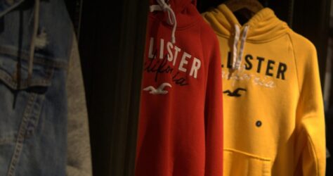 Red and yellow hoodies with the Hollister California logo hang on a clothing rack next to a gray denim jacket in a dimly lit store. BAM Agency