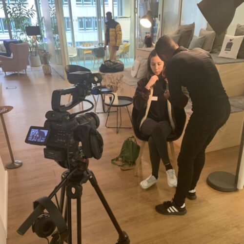 A person sits on a chair while another adjusts a microphone on their shirt, preparing for an interview. A video camera is set up in the foreground, and people are visible in the background of a bright indoor setting. BAM Agency