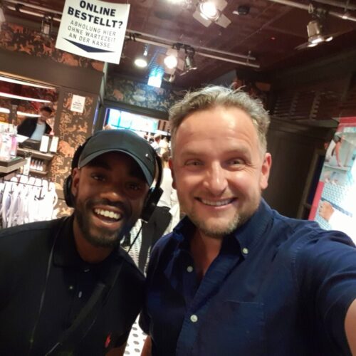 Two men smiling and posing for a selfie inside a brightly lit clothing store. There’s a sign in German above them and racks of clothes visible in the background. BAM Agency