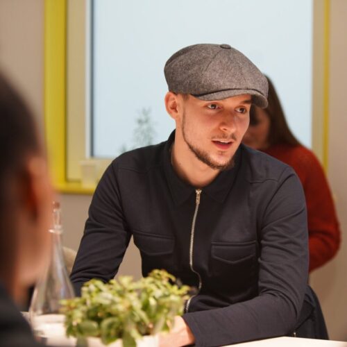 A young man with a trimmed beard, wearing a flat cap and a dark jacket, sits at a table with a green plant and water bottle, conversing in a bright room with yellow window trim. BAM Agency