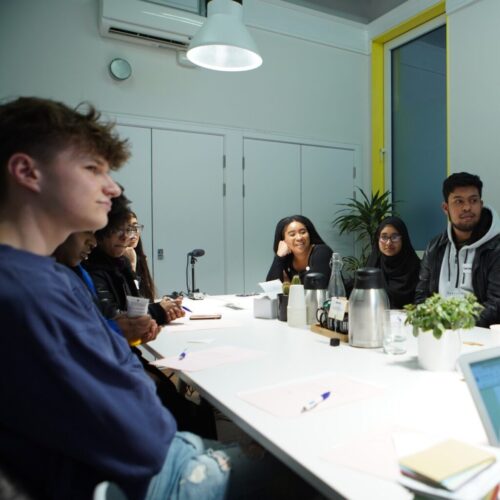 A group of young adults sit around a conference table in a modern meeting room, engaged in discussion. Papers, pens, and a laptop are on the table, with plants and a coffee pot also visible. Some people are smiling. BAM Agency