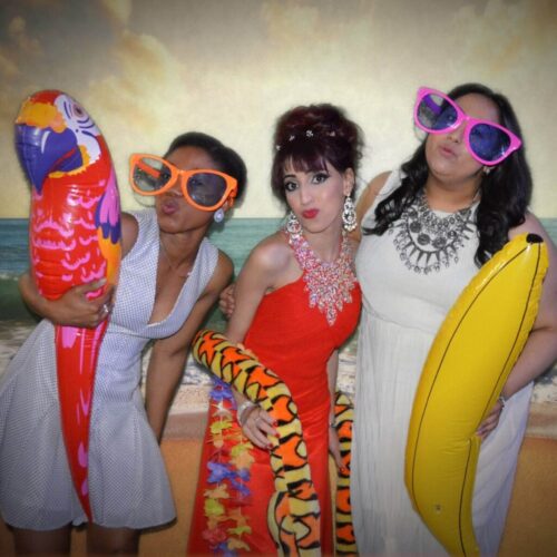 Three women posing playfully in front of a beach backdrop. They wear oversized sunglasses and hold inflatable props: a parrot, a snake, and a banana. One wears a red dress, the others in white and patterned dresses, all smiling. BAM Agency