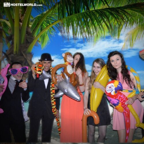 A group of five people having fun with inflatable toys and oversized accessories in front of a tropical beach backdrop. They are dressed in formal attire and are holding items like a parrot, monkey, and sunglasses. BAM Agency