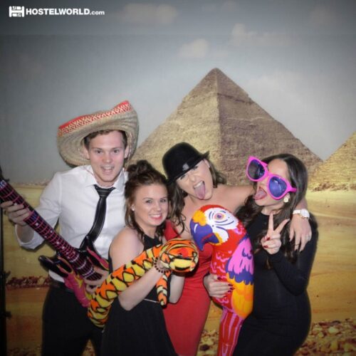 Four people pose in front of a backdrop featuring pyramids. They are dressed in playful attire, including a sombrero, oversized sunglasses, and inflatable animals. One person is playing an inflatable guitar, while others smile and gesture excitedly. BAM Agency
