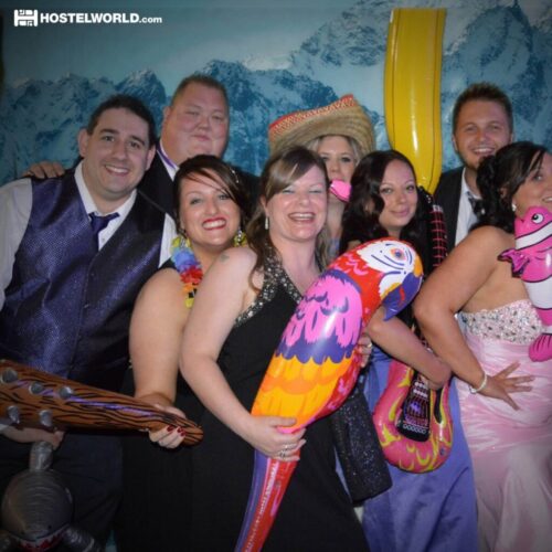A group of people in party attire, posing with inflatable props like a parrot, fish, and guitar. They are in front of a scenic backdrop featuring mountains and snow. The mood is festive and joyful. BAM Agency