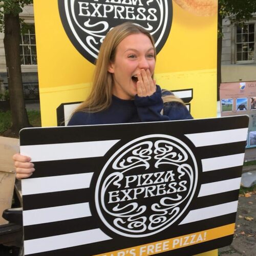BAM Agency A person stands outdoors, excitedly holding a large sign with the Pizza Express logo and text offering a years free pizza. Trees and a building are in the background. BAM Agency