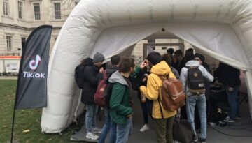 BAM Agency A group of people gathered outside a white inflatable structure on a grassy area. A black flag with the TikTok logo stands nearby, showcasing brand on campus initiatives. The background reveals a building with large windows and classical architecture. BAM Agency