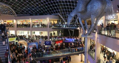BAM Agency Interior of a bustling shopping mall with multiple levels. A large, abstract horse sculpture hangs from the ceiling. Shoppers are seen throughout the levels, with stores and bright lights adding to the vibrant atmosphere. BAM Agency