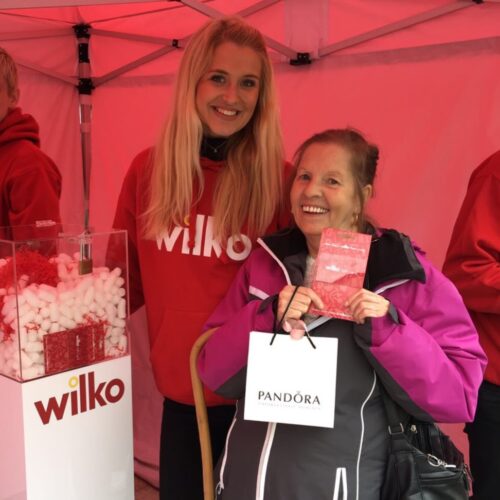 Two smiling women stand under a red tent. The woman on the left wears a red Wilko hoodie. The woman on the right holds a Pandora shopping bag and a small red packet. A Wilko-branded display with packing materials is visible behind them. BAM Agency