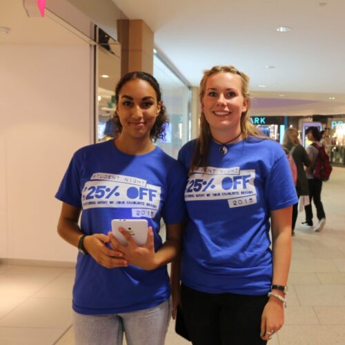 Two people stand in a shopping mall, smiling at the camera. Theyre wearing blue T-shirts emblazoned with 25% OFF, promoting a student night discount event by Land Securities. One holds a phone, and the other carries a bag, embodying the thrill of great deals. BAM Agency