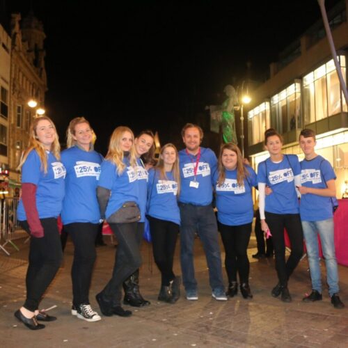 Nine people wearing blue shirts with 25% OFF printed on them pose together outdoors at night. Against the backdrop of Land Securities brilliantly illuminated buildings, they stand proudly, creating a striking scene in the lit area. BAM Agency