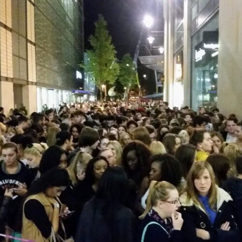 A large crowd gathered outside a Land Securities building at night, many looking towards the entrance. Some are on their phones, clad in casual attire. The scene is gently illuminated by streetlights and building lights. BAM Agency