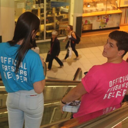 Two people wearing Official Freshers Helper shirts, one in blue and one in pink, ride an escalator. The person in pink holds a stack of flyers. In the background, Land Securities bustling shopping area is alive with pedestrians exploring their surroundings. BAM Agency