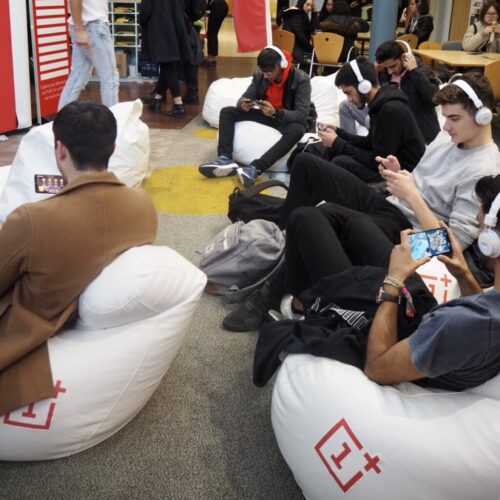 A group of people sitting on white bean bags, wearing headphones, and focused on their smartphones. They are in a public area with others in the background. The bean bags have a red 1+ logo. BAM Agency